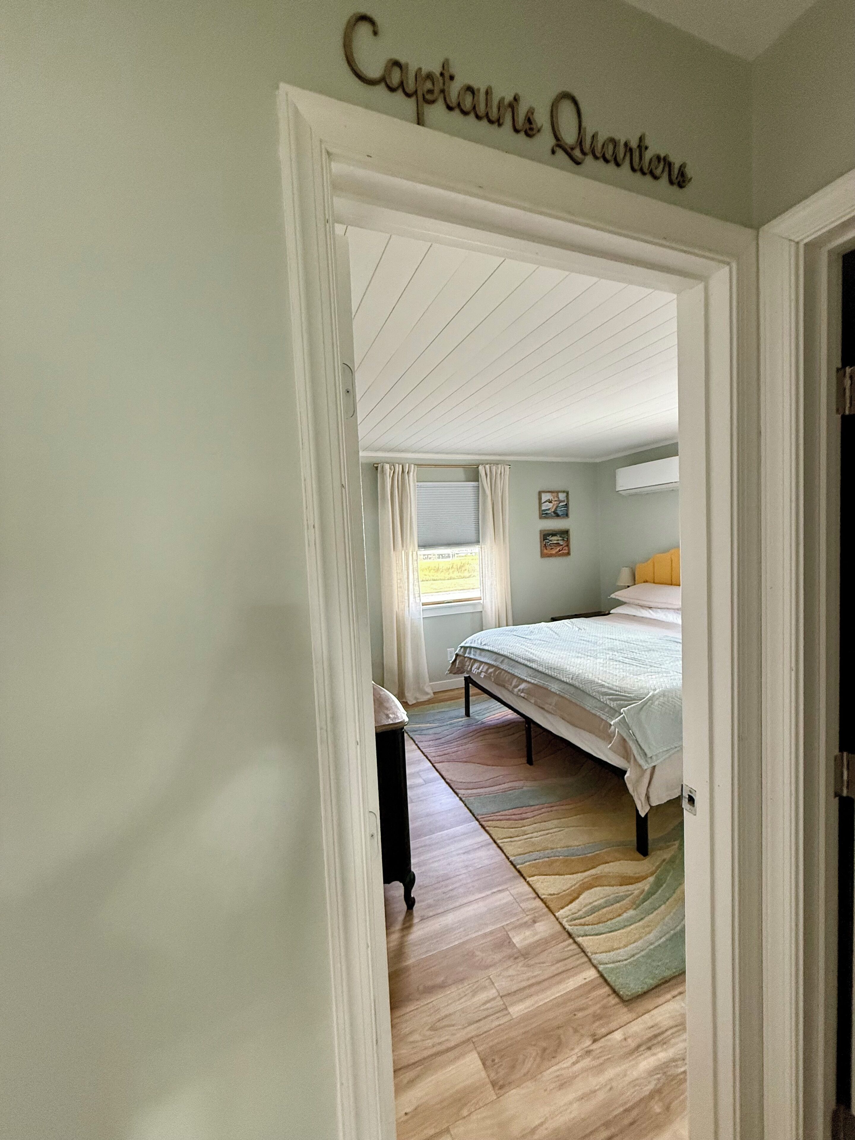 A bedroom with a bed, a window with curtains, a painting on the wall, and a sign above the door reading "Captains Quarters".