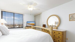Habitación de hotel con una cama grande, ventilador de techo, televisión, cómoda y ventana con vista a la ciudad.