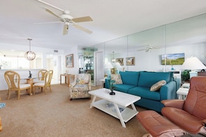 A spacious living room with a teal sofa, a brown leather armchair, and a white coffee table. There is a dining area with wooden chairs and a small table. A large mirror reflects the room, and a ceiling fan is visible.