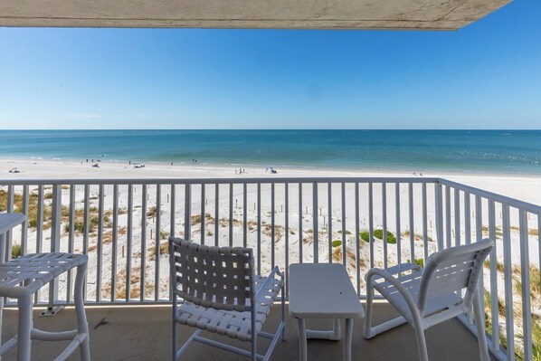 Ein Balkon mit weißen Stühlen und einem Tisch mit Blick auf einen Strand, an dem sich Menschen befinden und ein Volleyballnetz steht.