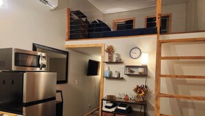A compact kitchen with a microwave, refrigerator, and wooden cabinets. A loft bedroom with a bed and a window is visible above the kitchen.