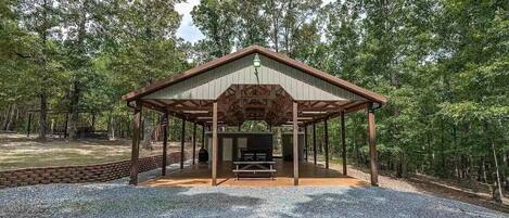 A covered pavilion with a picnic table and grill, surrounded by a forested area.