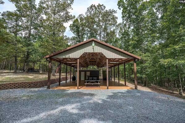 A covered pavilion with a picnic table and grill, surrounded by a forested area.