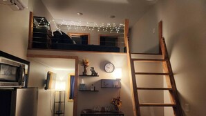 A loft-style room with a kitchen area, a wooden ladder, a clock, and string lights.