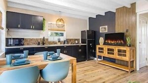 A modern kitchen with dark cabinets, a wooden dining table with blue chairs, a built-in TV, and a wooden cabinet with a TV stand. - Citylife Apartments - Pool / Free Parking / in Willemstad. Apartment I (Willemstad)