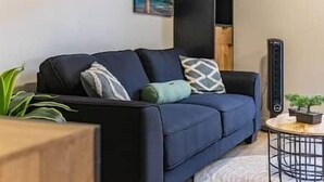 A living room with a dark blue sofa, a small round table, and a tall black radiator. - Citylife Apartments - Pool / Free Parking / in Willemstad. Apartment I (Willemstad)