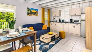 En kompakt opholdsstue med spiseplads, sofa, køkken med hvide skabe og et vindue med udsigt til grønne områder.