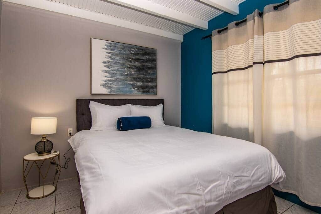 Un lit bien fait, avec du linge de lit blanc et un oreiller bleu, une lampe de chevet et un tableau accroché au mur.