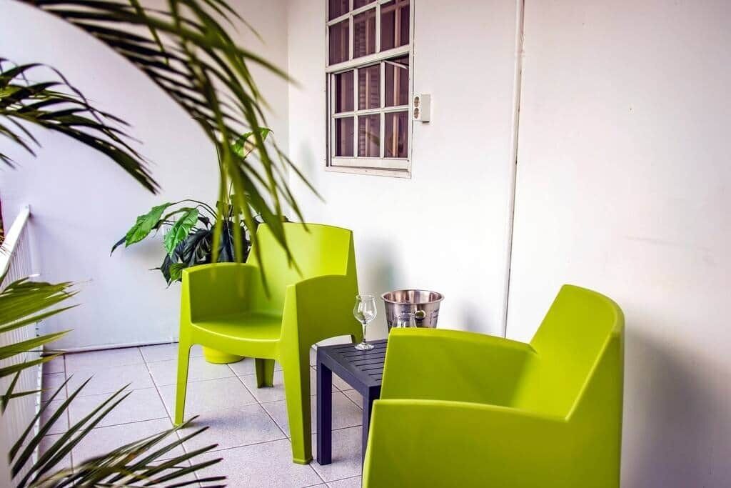 Un petit espace extérieur aménagé avec deux chaises vert vif et une table, un pot de fleurs et une fenêtre ornée d’une grille métallique.
