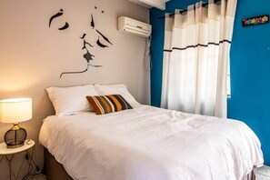 Un dormitorio con cama, aire acondicionado de pared, lámpara de noche y ventana con cortinas.