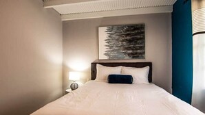 Ein ordentlich bezogenes Bett mit weißen Leinen und einem dunklen Kopfteil, eine Nachttischlampe und ein gerahmtes Kunstwerk darüber.