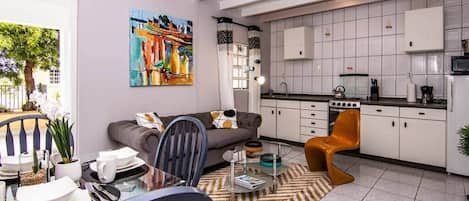 A modern kitchen with white cabinets, a dining area with a glass table, and a living room with a sofa and a painting.