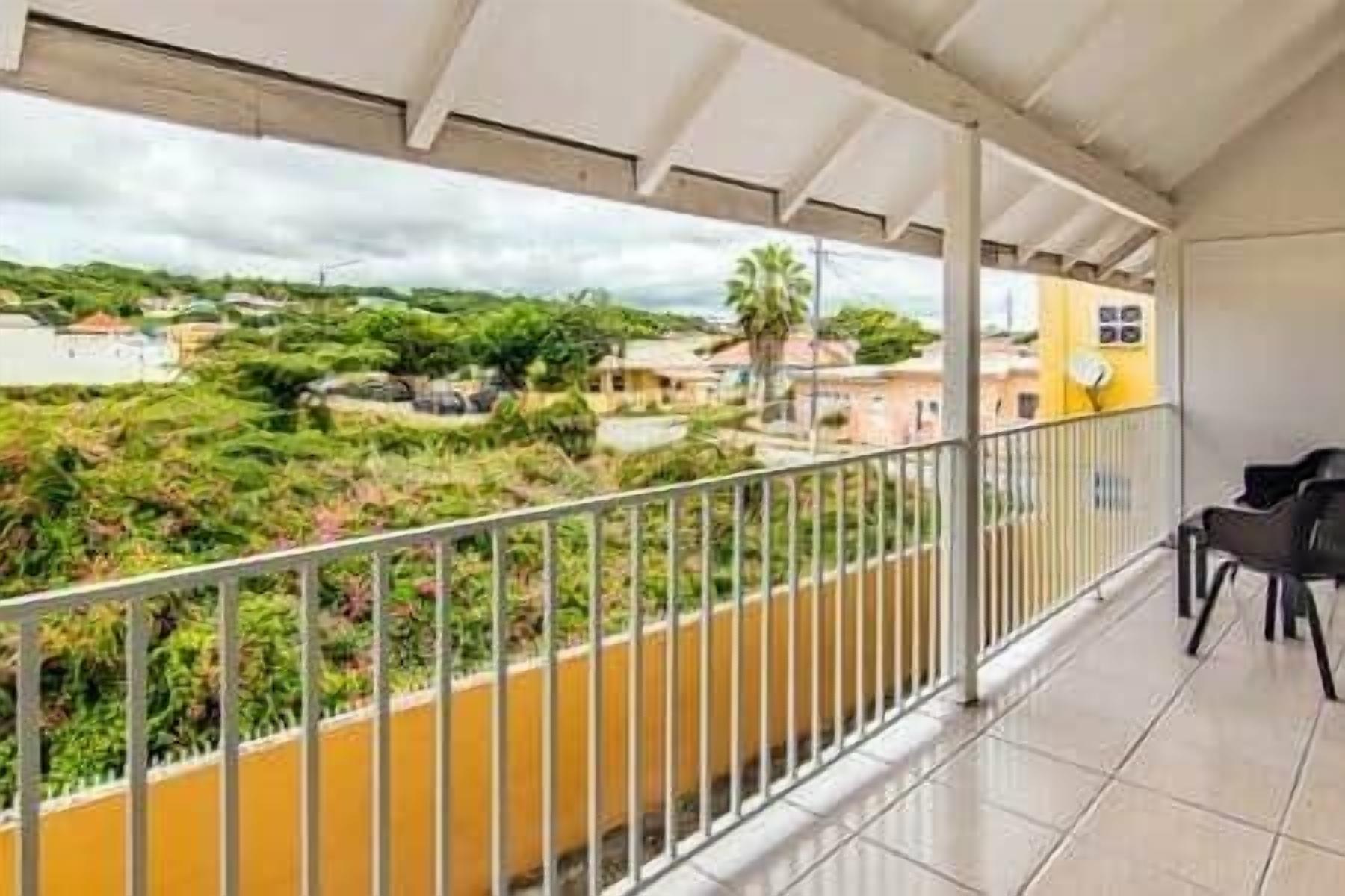 A balcony with a metal railing, a yellow wall, and a view of a residential area with houses and greenery.