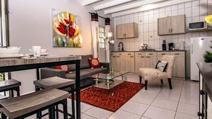 Una cucina moderna con mobili bianchi, un forno a microonde e un frigorifero. C'è una zona pranzo con un tavolo in vetro e sedie, e un divano con cuscini. Un quadro astratto e colorato adorna la parete.