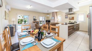A dining area with a table set for four, a kitchen with modern appliances, and a view of the outdoors through large windows. - Villa Alexander - Three Bedroom Villa, Sleeps 6 (Paralimni)