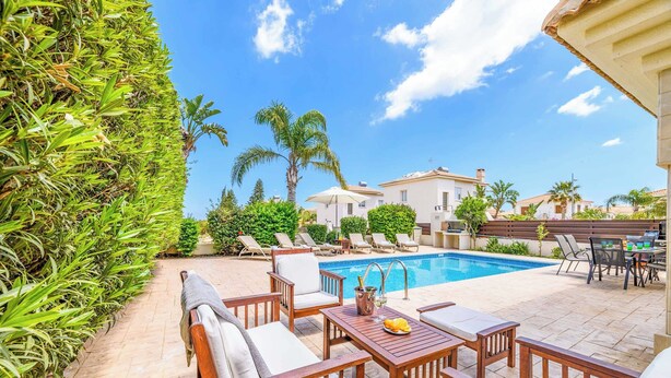 A poolside area with wooden furniture, a pool, and palm trees. - Villa Alexander - Three Bedroom Villa, Sleeps 6 (Paralimni)