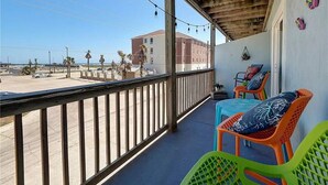 Un balcon doté d’une balustrade en bois, deux chaises en plastique de couleurs vives, une table et une vue sur la plage et les bâtiments au loin.