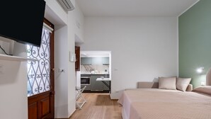 Une chambre avec un lit, un téléviseur fixé au mur, une cuisine à l’arrière-plan et une porte ornée d’un panneau métallique décoratif.