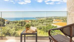 Un balcon avec vue sur la mer et la verdure, une chaise en osier et une table avec des livres.