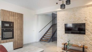Un salon moderne comprenant un canapé, une table basse, un téléviseur fixé au mur en pierre, un escalier en bois et des appareils électroménagers intégrés.