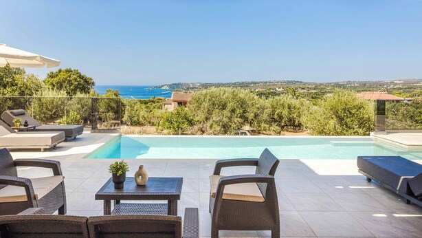 Un área de piscina en la azotea con sillones reclinables y una mesa, con vistas al mar azul turquesa y vegetación exuberante.
