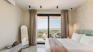 Habitación de hotel con una cama grande, un banco con objetos encima, un espejo y vista al mar a través de un ventanal amplio.