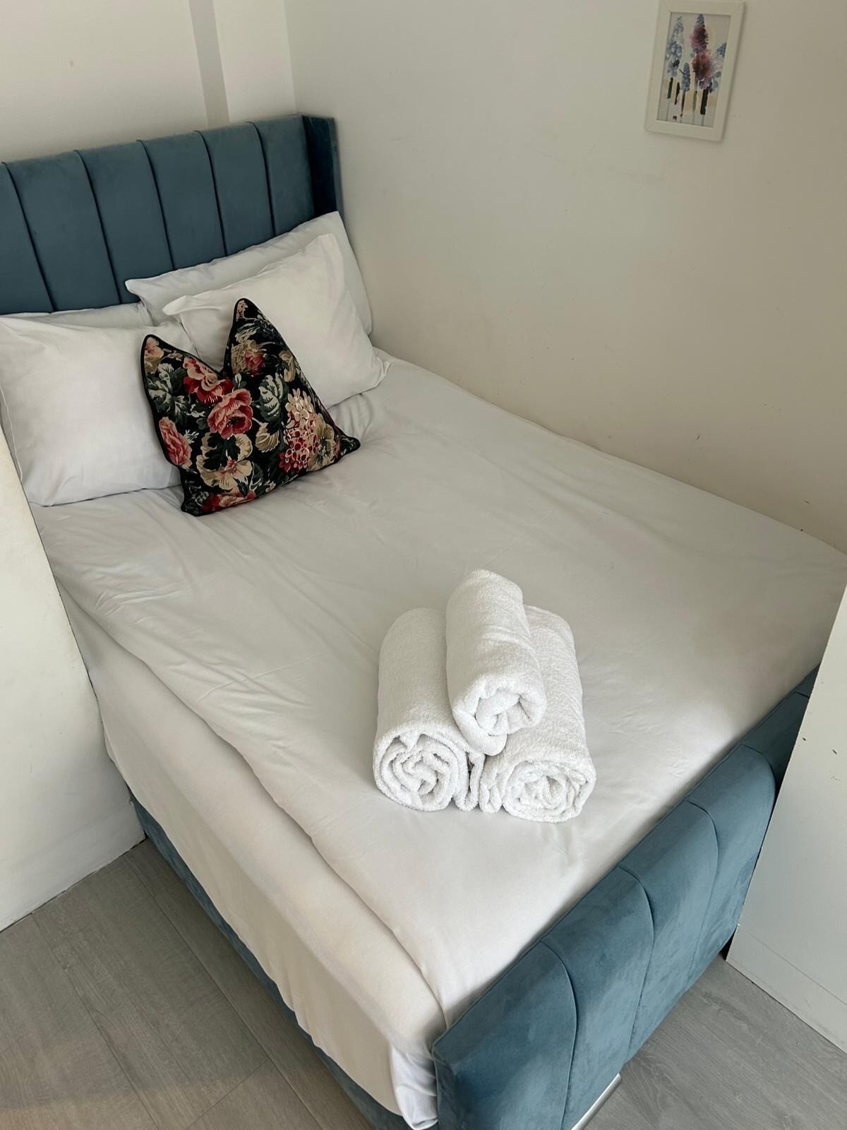 A neatly made bed with white linens and two rolled towels, a floral pillow, and a blue headboard.