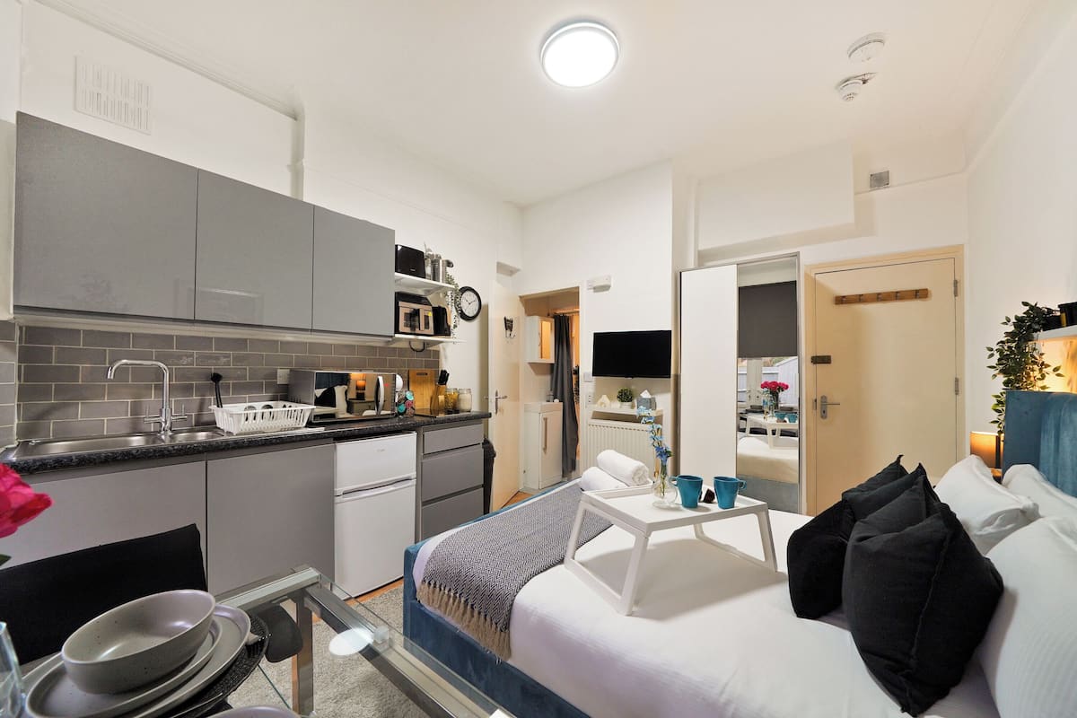 Une chambre d’hôtel compacte avec une kitchenette, un lit avec une literie noire et blanche, et une petite table avec une tasse bleue.