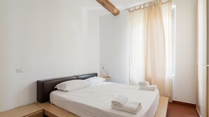 Un dormitorio con cama, mesita de noche, ventana con cortinas y viga de techo de madera.