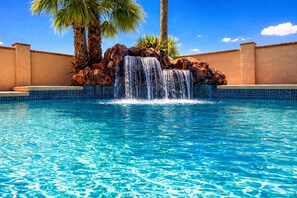 Una piscina azul cristalina con una cascada, rodeada de una pared de azulejos y palmeras.