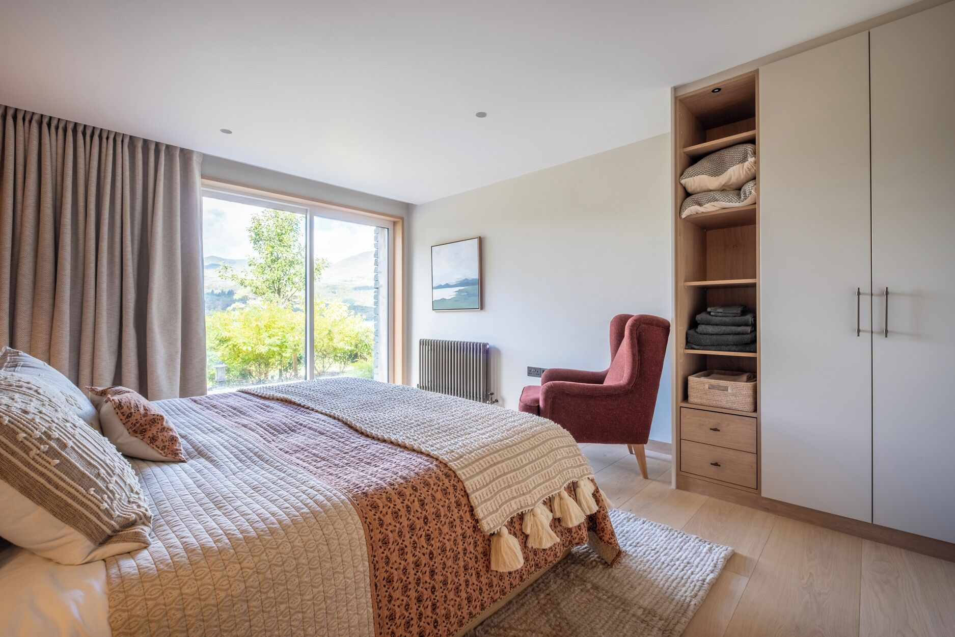 A bedroom with a large bed, a red chair, a wardrobe, and a window with a view of greenery.