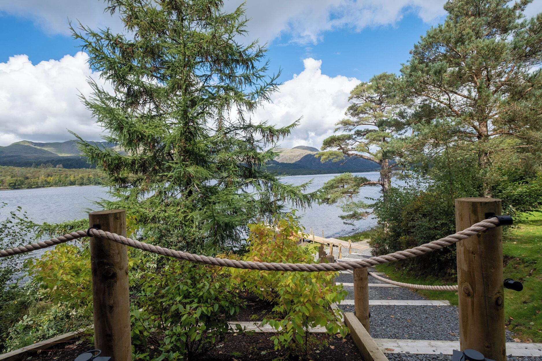 Ein Holzweg führt zu einem See, umgeben von Bäumen und Bergen, mit einer Seilbarriere.