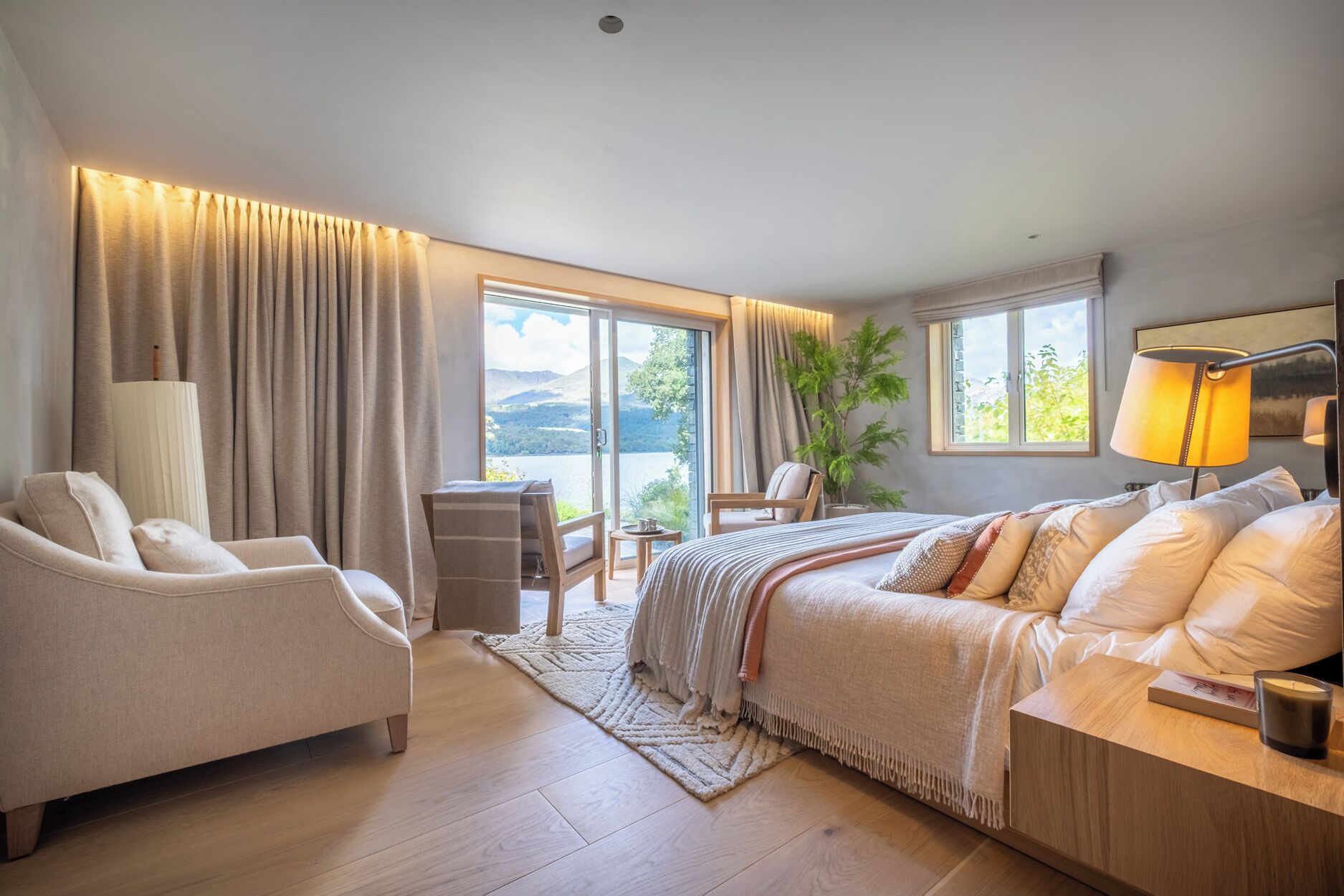 Un dormitorio amplio con una cama grande, un espacio de descanso con una silla y una mesa, y vistas a un lago a través de las ventanas.