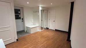 Um quarto amplo, com assoalho de madeira, paredes brancas e uma porta que dá para outro cômodo.