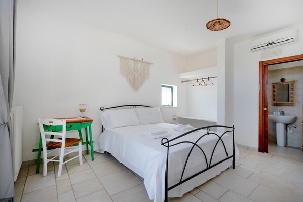 A bedroom with a bed, a desk, a chair, a hanging macrame, a window, and a bathroom with a sink.
