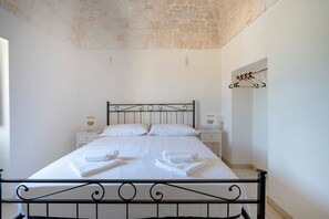 En pænt redt seng med hvide sengetæpper og håndklæder, et stenspecifikt loft og et simpelt natbord med en lampe.