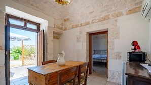 Una habitación rústica con una mesa de madera, sillas, una jarrón blanco y una cama al fondo.