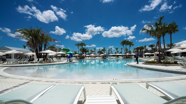 A large outdoor swimming pool with multiple lounge chairs and umbrellas, surrounded by palm trees and shaded areas. - Super Modern 7BR Windsor Cay* (Clermont)