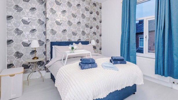 A bedroom with a white bed, blue curtains, and a white bedside table. - Sleep 7, Pool Table/TTennis, Free Parking, 3BR (Manchester)