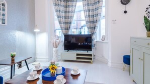 A modern living room with a television, a dining table with cups and saucers, a blue ottoman, and a cabinet. - Sleep 7, Pool Table/TTennis, Free Parking, 3BR (Manchester)
