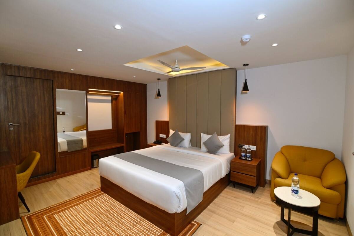 A modern hotel room with a large bed, wooden wardrobe, a mirror, a desk with a chair, and a small round table with a bottle.