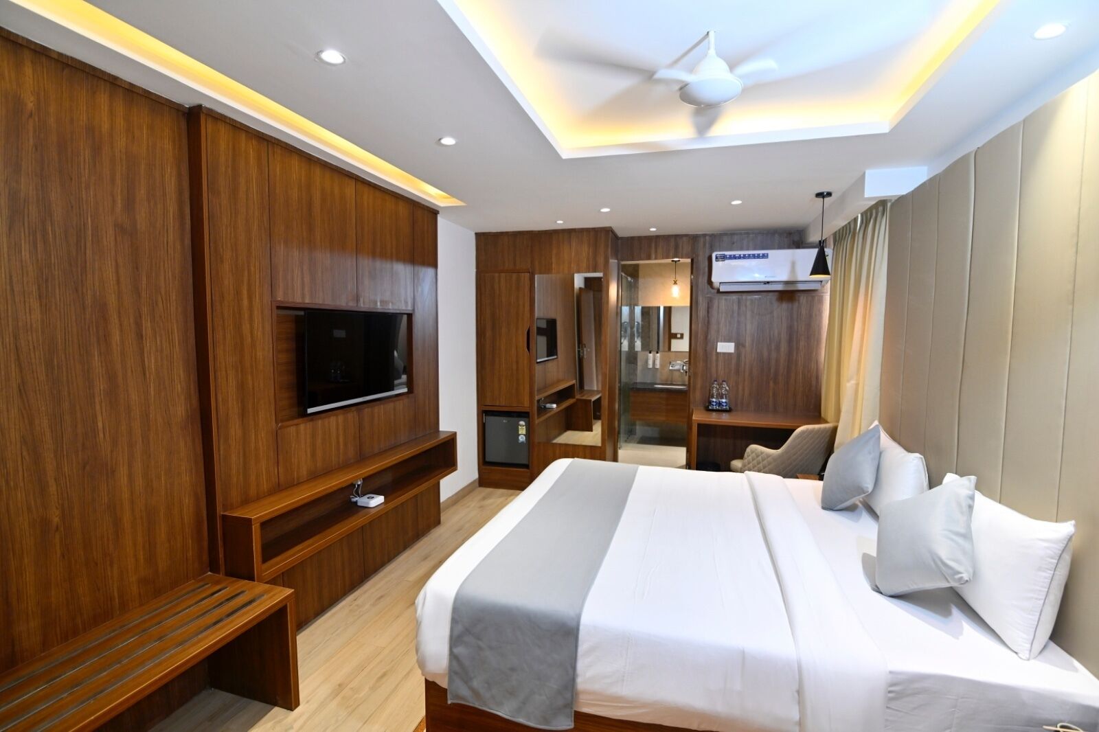 A modern hotel room with a large bed, wooden paneling, a flat-screen TV, a ceiling fan, and a small kitchenette.
