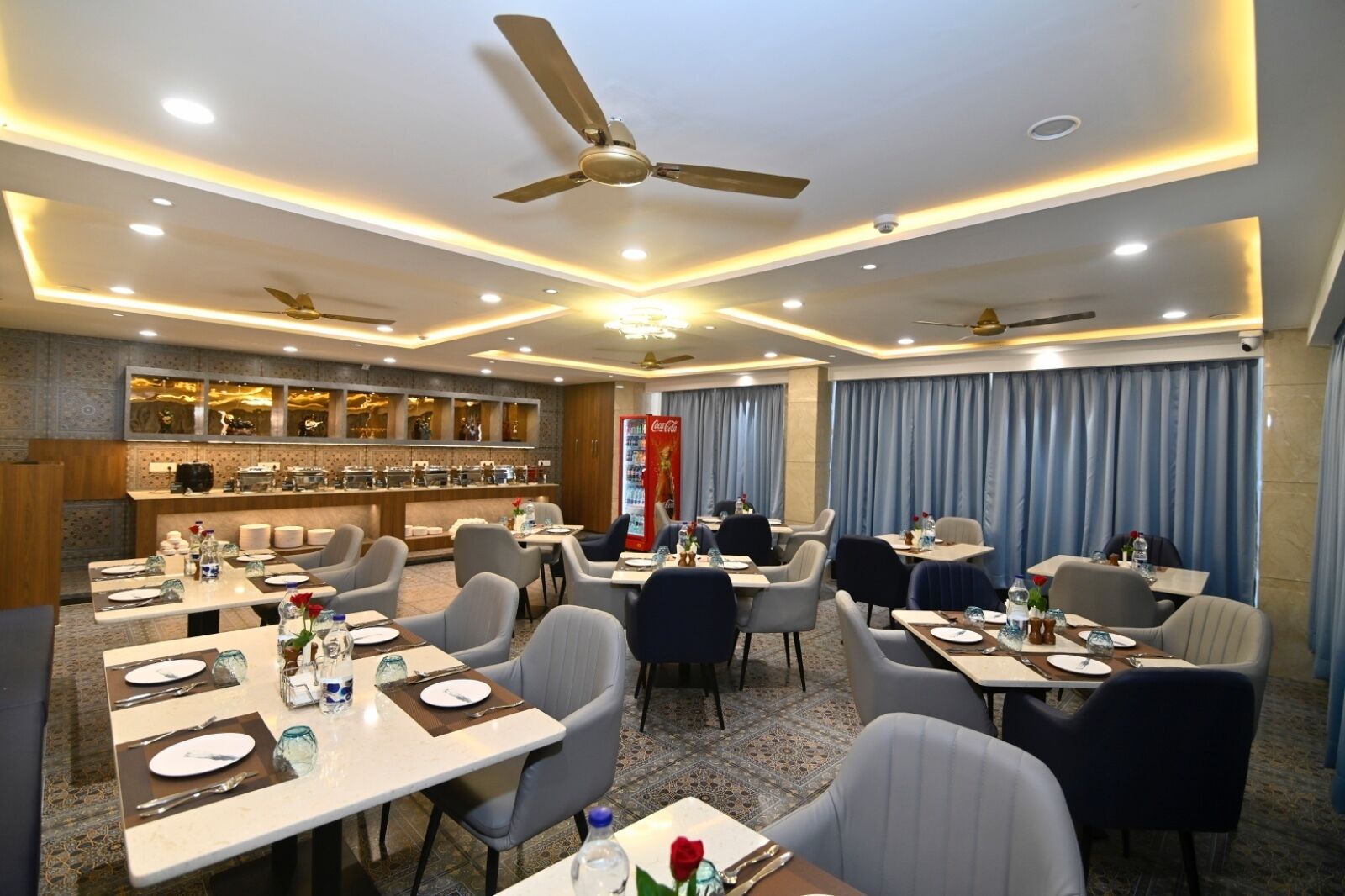 Un comedor de hotel con varias mesas, sillas, ventiladores de techo y una barra de buffet.