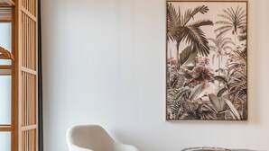 A bedroom with a bed, a white chair, a wooden crib, and a tropical-themed wall art.