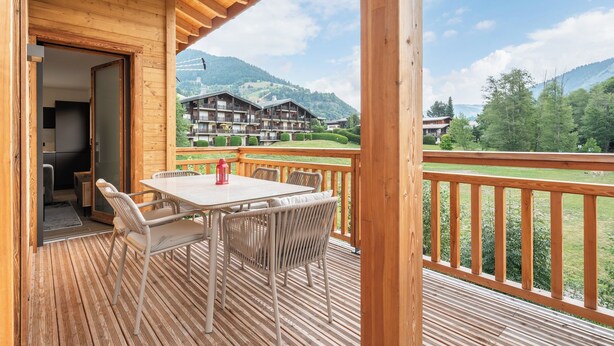 A wooden deck with a table and chairs, overlooking a mountainous landscape and a hotel building.