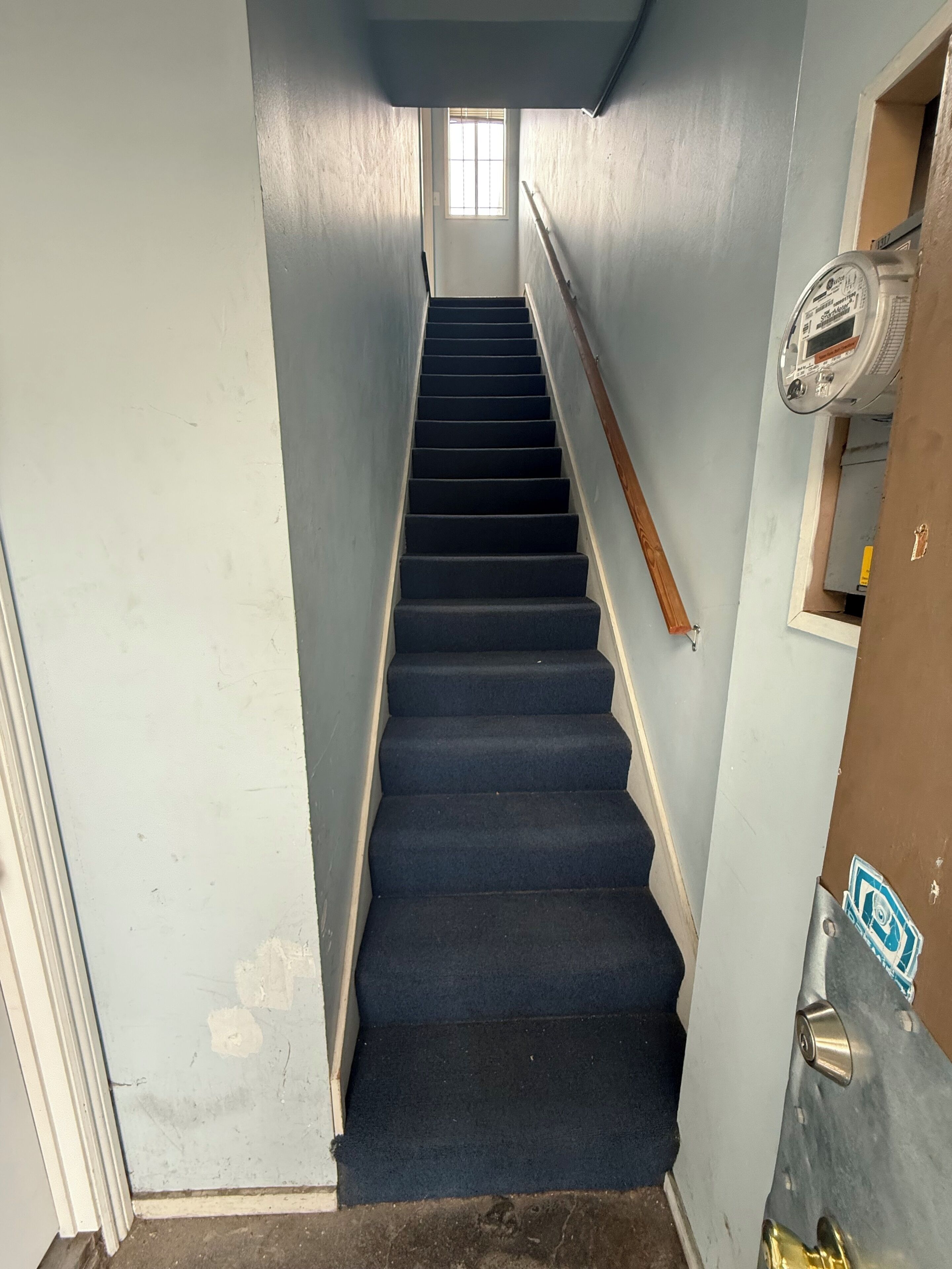 A narrow stairway with blue carpet leading to a door with a thermostat and a lock.