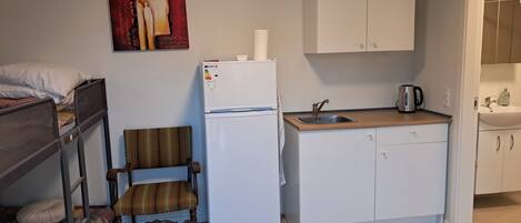 Una kitchenette compacta con refrigerador, lavamanos y una silla de madera.