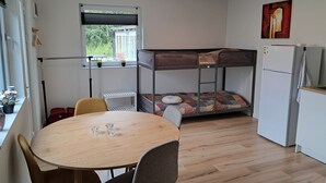 Una habitación compacta con literas, una pequeña cocina con refrigerador, una mesa de comedor con sillas y un ventanal con vista a la vegetación.