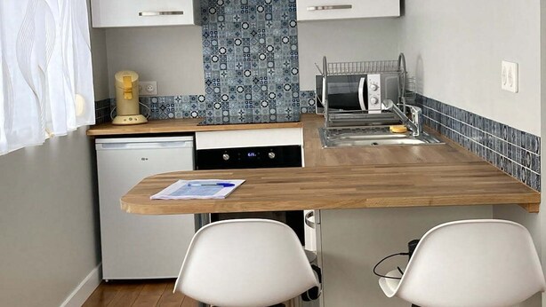 Ett kompakt kök med vita skåp, en blå, mönstrad väggbeklädnad bakom arbetsbänken, en liten diskmaskin och en köksö i trä med två barstolar.