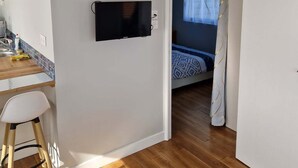Ett rum med trägolv, en säng med ett mönstrat överkast, en tv som är monterad på väggen, en liten köksdel med ett handfat och en stol samt en dörr som leder till ett annat rum.